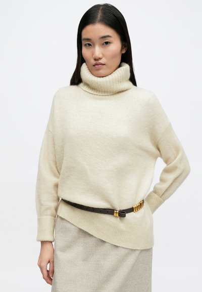 Κρεμ ζακέτα με πλεχτό τούλπες, χαρακτηρισμένη από χαλαρή εφαρμογή και ριγέ γιακά, συνδυασμένη με μία καφέ δερμάτινη ζώνη με χρυσές λεπτομέρειες.