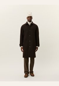 Brown wool overcoat with large buttons, side pockets, and a collar, worn over a white turtleneck and beige trousers, paired with brown shoes.
