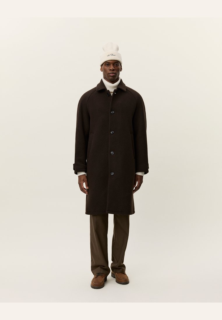 Brown wool overcoat with large buttons, side pockets, and a collar, worn over a white turtleneck and beige trousers, paired with brown shoes.