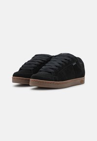 Sorte semskede sneakers med gummisåler, snøredesign og kontrasterende brun såle. Har teksturerte detaljer og minimal merkevare.