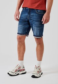 Shorts de mezclilla en azul oscuro con detalles desgastados, que presentan un ajuste slim y dobladillos turnados. Combinados con zapatillas blancas y con estampado.