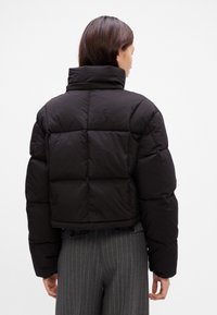 Femme vue de dos portant une doudoune noire à col haut et un pantalon gris à fines rayures sur un fond uni.