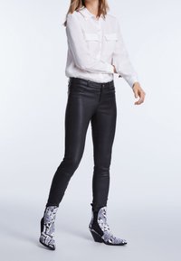 Femme portant une chemise blanche boutonnée, un pantalon en cuir noir et des bottines à talons à motif peau de serpent noir et blanc, debout devant un fond uni.
