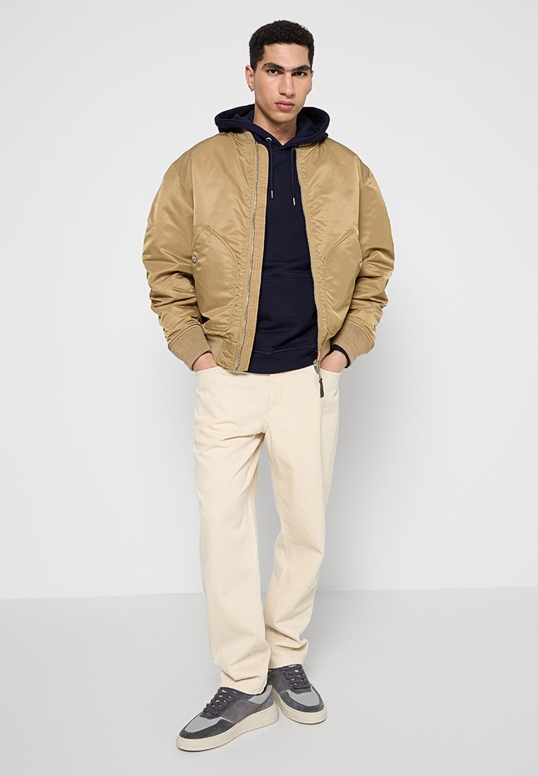 Giacca bomber tan con finitura testurizzata, felpa con cappuccio blu navy, pantaloni crema e sneakers grigie, con accenti bianchi e scuri.