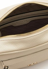 Interno di una borsa in pelle testurizzata color crema con cerniera dorata, dotato di una fodera a motivo scuro e un logo impresso sul lato.