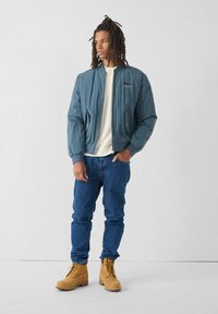 Giacca bomber azzurro chiaro con polsini a maglia, indossata sopra una camicia crema, abbinata a jeans blu e stivali con lacci marroni.