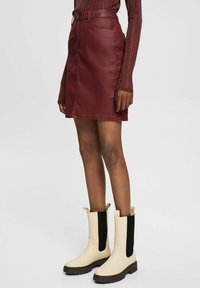 Femme portant une jupe en cuir bordeaux, un haut à manches longues assorti et des bottes crème avec des panneaux latéraux élastiques noirs.