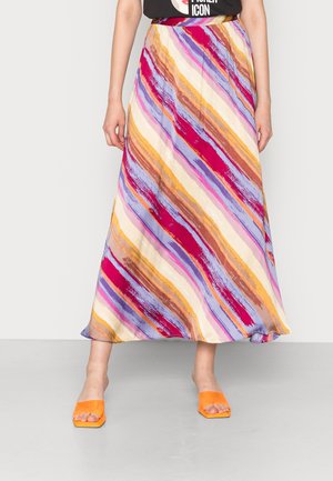 Femme portant une jupe midi rayée diagonale colorée avec des nuances de rouge, violet, jaune et marron, assortie à des sandales orange à bout ouvert.