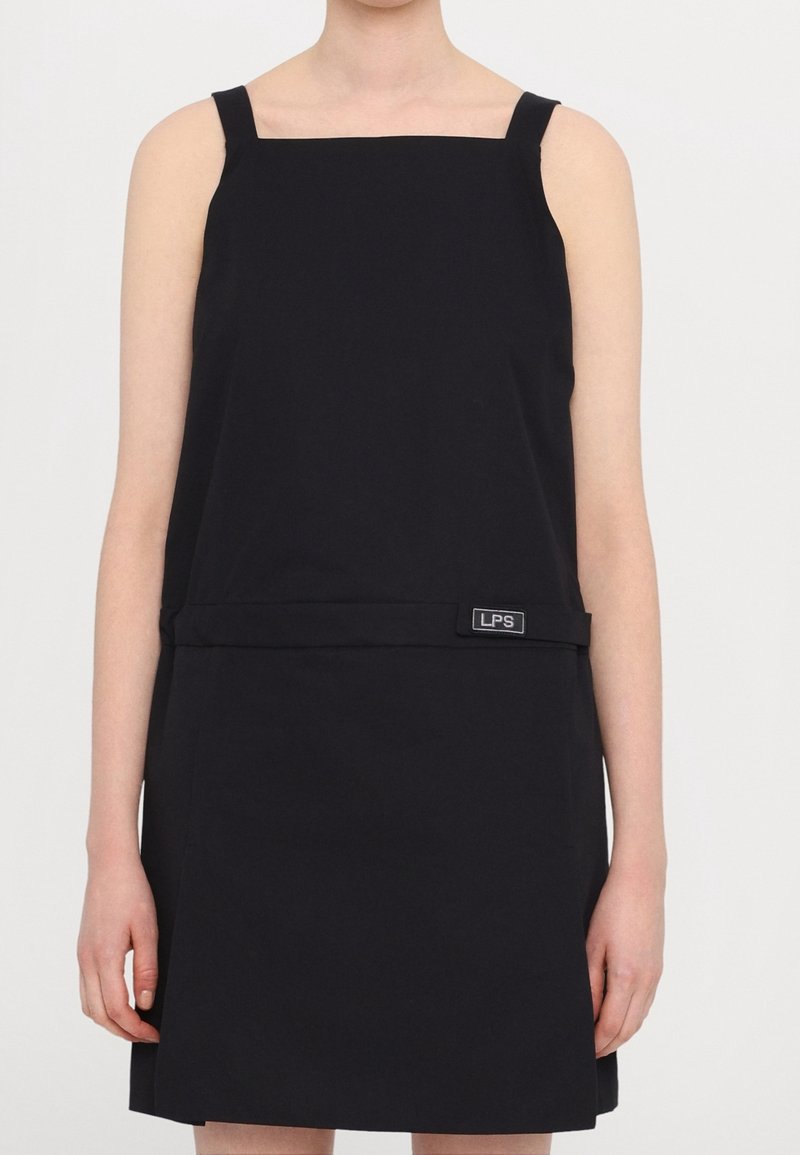 Vestido negro sin mangas con escote cuadrado, hecho de una tela suave. Presenta una banda horizontal y un logo en la cintura. Forma A-line sencilla.