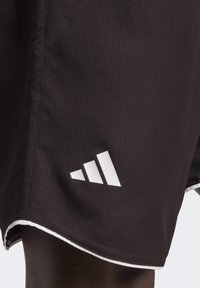 Juodi sportiniai šortai su grioveliais, balta apdaila ir ryškiu baltu trefoil logo apatinėje kairėje pusėje.