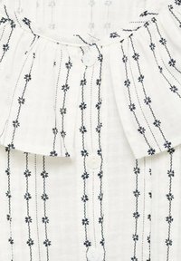 Camicia bianca con colletto a volant, caratterizzata da ricami floreali navy e chiusura con bottoni. Tessuto testurizzato con linee verticali e motivi.