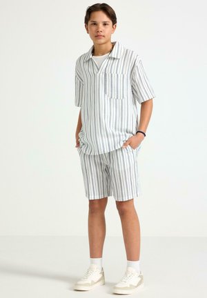 Teenager iført hvid t-shirt med korte ærmer og shorts med lodrette grå striber, hvide sokker og beige sneakers, står med hænderne i lommerne.