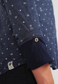 Donkerblauwe langemouwenhemd met witte kleine weer- en natuuriconen, met opgerolde mouwen die vastgezet zijn met een houten knoop.