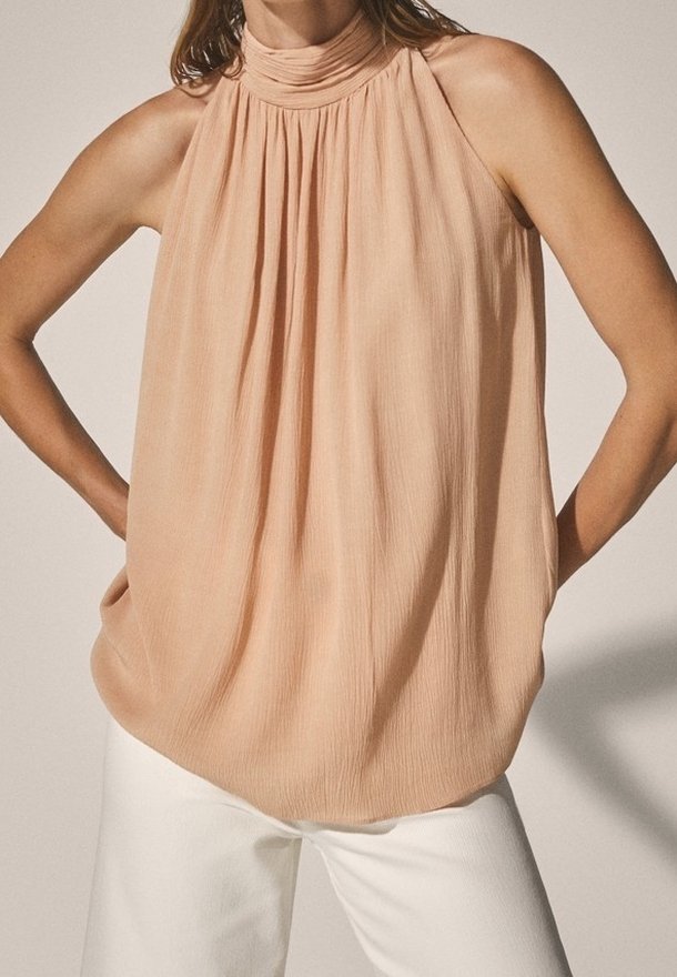 Femme portant un chemisier sans manches couleur pêche à col montant, associé à un pantalon blanc, bras pliés et mains posées sur les hanches.