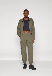 Chaqueta corta de color verde oliva con cremallera frontal y puños acanalados, combinada con pantalones cargo a juego que incluyen bolsillos y puños elásticos. Zapatillas deportivas negras.