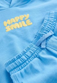 Světle modrá mikina s kapucí s žlutým potiskem "HAPPY SMILE" na přední straně, doplněná o odpovídající tepláky s elastickým pasem a stahovací šňůrkou.