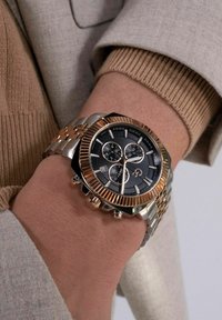 Metāla rokas pulkstenis ar melnu ciparnīcu, rozā zelta akcentiem un teksturētu rāmīti. Sudraba un zelta saite, kas ietver trīs apakšciparnīcas.