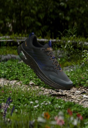 Zwart-oranje trailschoen met een gespikkelde zool, zwevend boven een grasachtig, rotsachtig pad in een bosrijke omgeving.