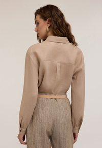 Beige linne skjorta med krage och långa ärmar, tillsammans med ljusbruna randiga byxor och ett tunt beigebälte. Texturerade tygdetaljer synliga.