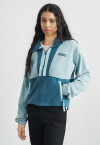 Νεαρή γυναίκα με μακριά σκούρα μαλλιά που φοράει μπλε και τυρκουάζ φλις ζακέτα Columbia με φερμουάρ και μαύρο παντελόνι, στέκεται μπροστά σε λευκό φόντο.