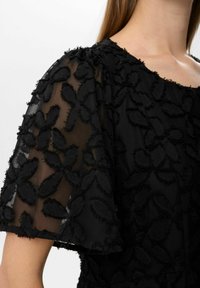 Rochie neagră cu mâneci transparente, având aplicații florale texturate și margini desfăcute. Țesătură netedă cu o siluetă ajustată.