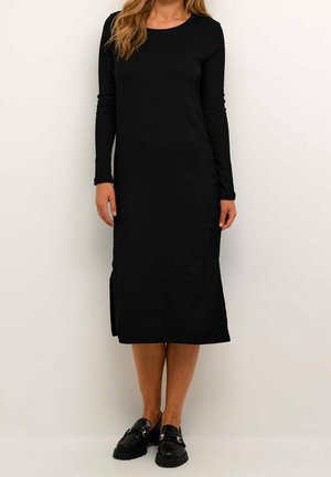 Kvinde stående iført en sort midi-kjole med lange ærmer og sideslid samt sorte loafers mod en ensartet lys baggrund.