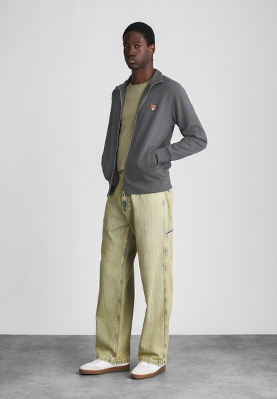 Gray zip-up jacket with pockets, green t-shirt underneath, and pale yellow baggy jeans. White sneakers with brown soles complete the outfit.