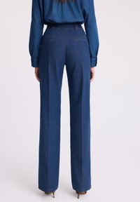 Kobieta widoczna od tyłu, ubrana w ciemnoniebieskie spodnie dżinsowe o wysokim stanie z prostymi nogawkami oraz dopasowaną, długą koszulę dżinsową.