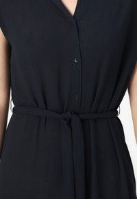 Vestito blu navy, a maniche corte, realizzato in un tessuto leggero e testurizzato. Presenta un scollo a V, chiusura con bottoni e una cintura annodata in vita.