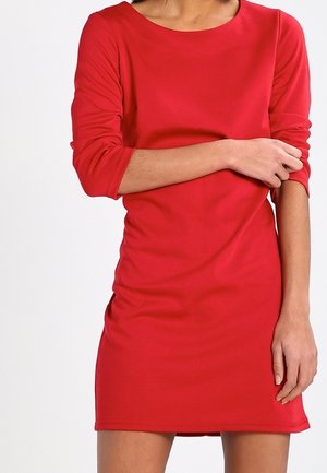Femme portant une robe rouge ajustée à manches longues, avec un bras plié tenant le coude opposé.
