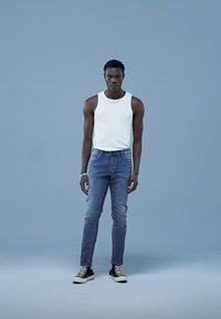 Bijela majica bez rukava, plave traperice, crne visoke tenisice. Model stoji ispred svijetloplave pozadine. Jednostavni nakit kao detalj.