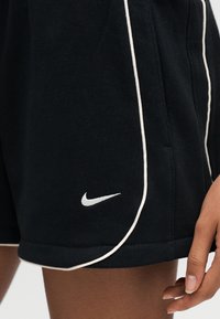 Μαύρο αθλητικό σορτς με λευκή διακόσμηση και λευκό λογότυπο Nike στο αριστερό πόδι, φορεμένο από άτομο με ορατό χέρι και σκούρο τόνο δέρματος.