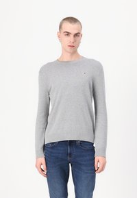 Grijze gebreide trui met lange mouwen, ronde hals, geribbelde zoom en klein logo-accent op de borst. Gecombineerd met blauwe spijkerbroek.
