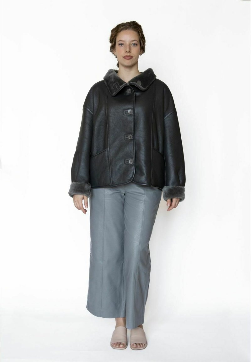 Zwarte leren jas met een brede kraag, vijf knopen en grijze bontaccenten. Gecombineerd met lichtgrijze leren broek en neutrale sandalen.