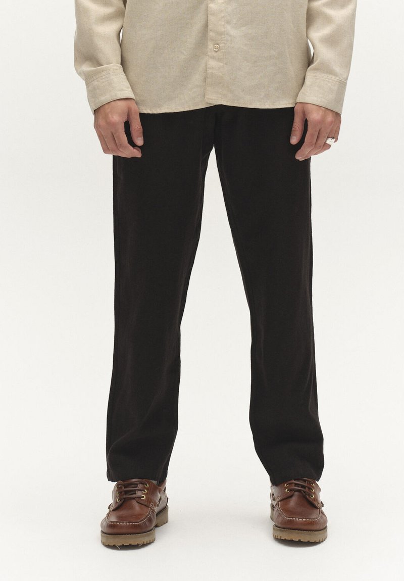 Muškarac u crnim hlačama, bež dugoj košulji i smeđim kožnim cipelama s vezicama stoji na jednobojnoj pozadini.