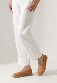 Persoon die witte linnen broek en beige gevlochten leren platte schoenen met gevlochten zolen draagt, staande op een lichte achtergrond.