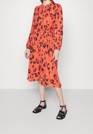 Femme portant une robe orange à manches longues avec motif léopard noir et marron, associée à des sandales noires à bout ouvert, debout devant un fond uni.