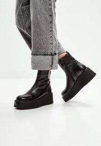 Černé kotníkové boty s hladkým koženým povrchem, které mají robustní platformu. Design zahrnuje boční zip a minimální detaily prošívání.
