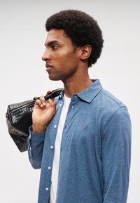 Camisa azul com botões, feita de tecido texturizado, usada sobre uma camisa branca. Bolsa preta de tecido trançado segurada por uma pessoa com cabelo curto e encaracolado.