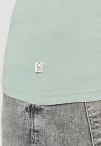Licht mintgroen T-shirt gemaakt van zachte stof, met een klein QS-label aan de zoom, gecombineerd met grijze stoffen jeans die details van textuur tonen.