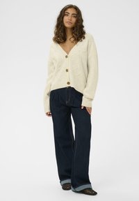 Krem kardigan s dugim rukavima i V-izrezom s ukrasnim uzorkom spirale, s smeškastim dugmadima, u kombinaciji s hlačama od tamnog denima visokog struka.