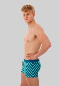 Shorts de bain avec un motif de rayures diagonales teal et bleu clair, fabriqués dans un matériau extensible avec une taille  élastique et un cordon de serrage.