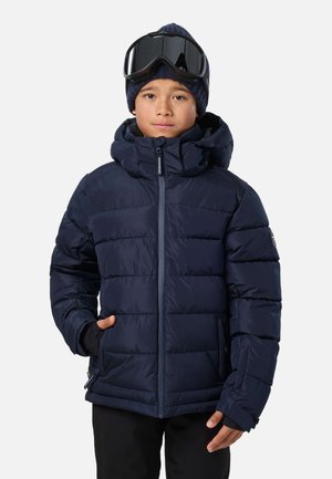 Garçon portant une doudoune bleu marine, un pantalon noir, un bonnet en laine bleu et un masque de ski noir sur le front, debout avec une main dans la poche.