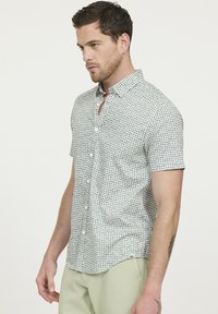 Chemise à manches courtes avec boutons, de couleur blanche, ornée d'un motif géométrique vert et noir. Fabriquée en tissu léger avec un col classique et une poche poitrine.