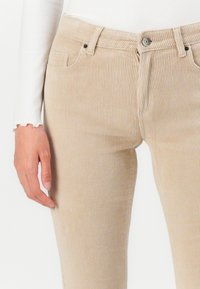 Beige corduroy pants with vertical ribbing, featuring front pockets, a button closure, and a zip fly. The fabric has a soft, textured appearance.