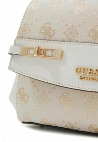 Sac à main en similicuir beige avec une fermeture à clapet dorée et un motif de logo embossé, doté d'une texture lisse et d'une forme structurée.