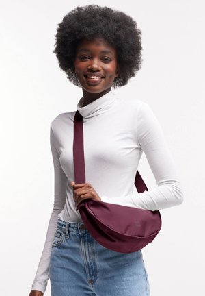 Bordeauxfarbene Schultertasche aus einem weichen Stoff. Runde Form mit einem breiten Tragegurt, gehalten von einer Person, die einen weißen Rollkragenpullover trägt.