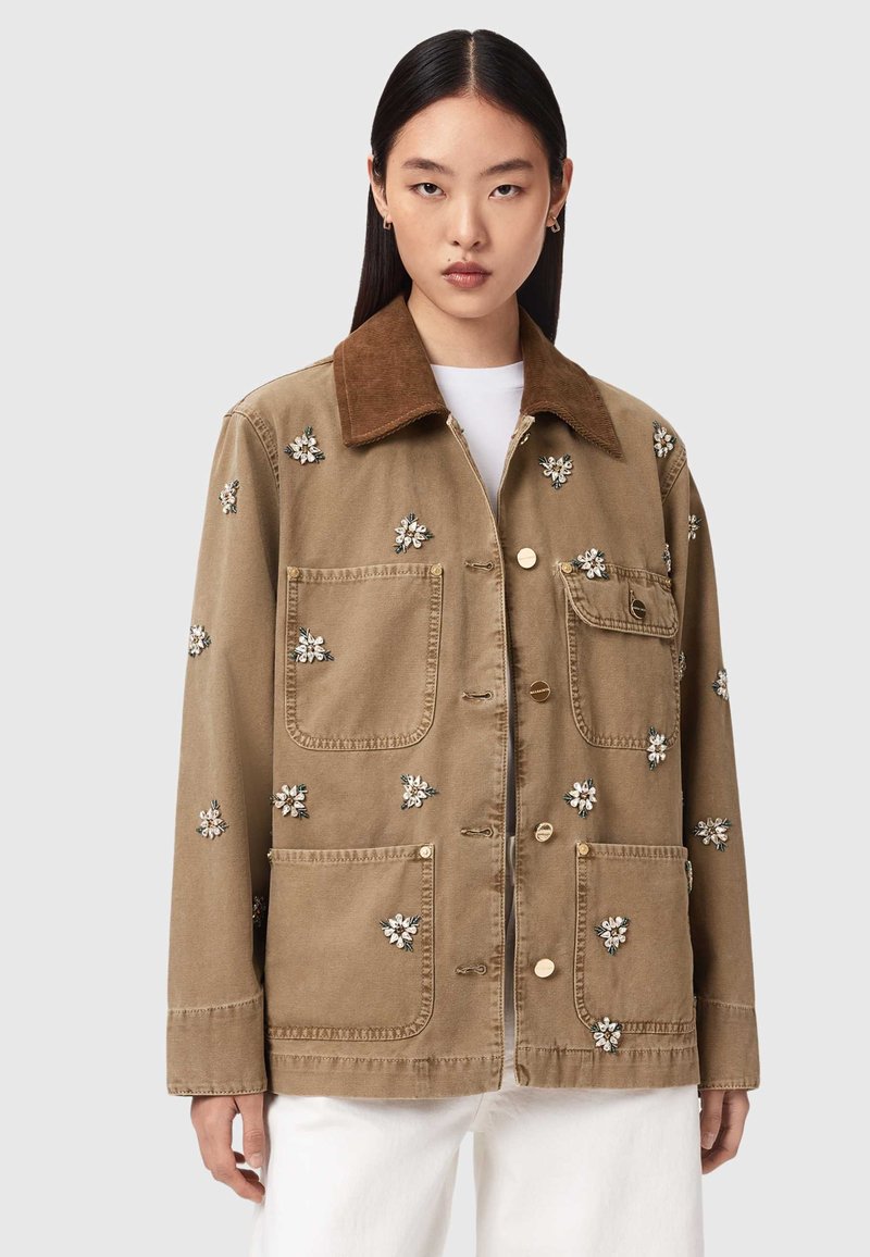 Frau trägt eine braune Jacke mit dunklerem Kragen und kleiner weißer Blumenstickerei, kombiniert mit weißen Hosen, vor einfachem Hintergrund.