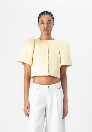 Top giallo corto con maniche a sbuffo e apertura frontale. Indossato con pantaloni bianchi a vita alta. Texture liscia, design minimal e stile aderente.