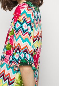 Blusa colorata con un motivo a zigzag in rosa, turchese e arancione, con accenti floreali e maniche a volant. Tessuto leggero.